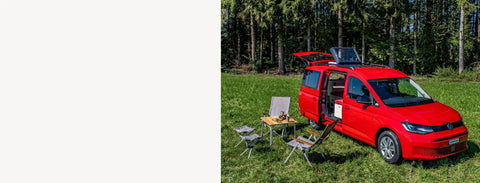 Roter VW-Minicamper mit geöffnetem Aufstelldach und Heckklappe auf einer Wiese am Waldrand, davor Campingtisch und Campingstühle